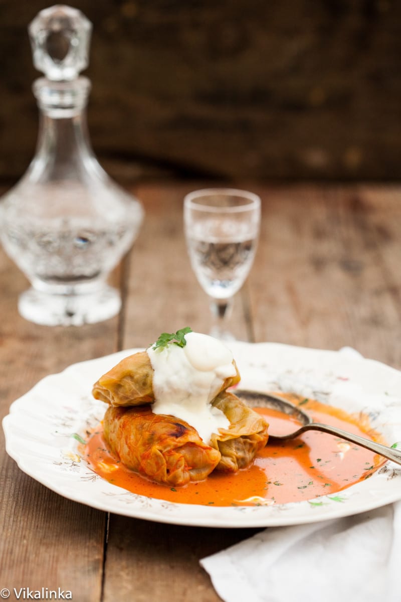 three cabbage rolls in sauce on a plate, decanter with clear liquid in the background