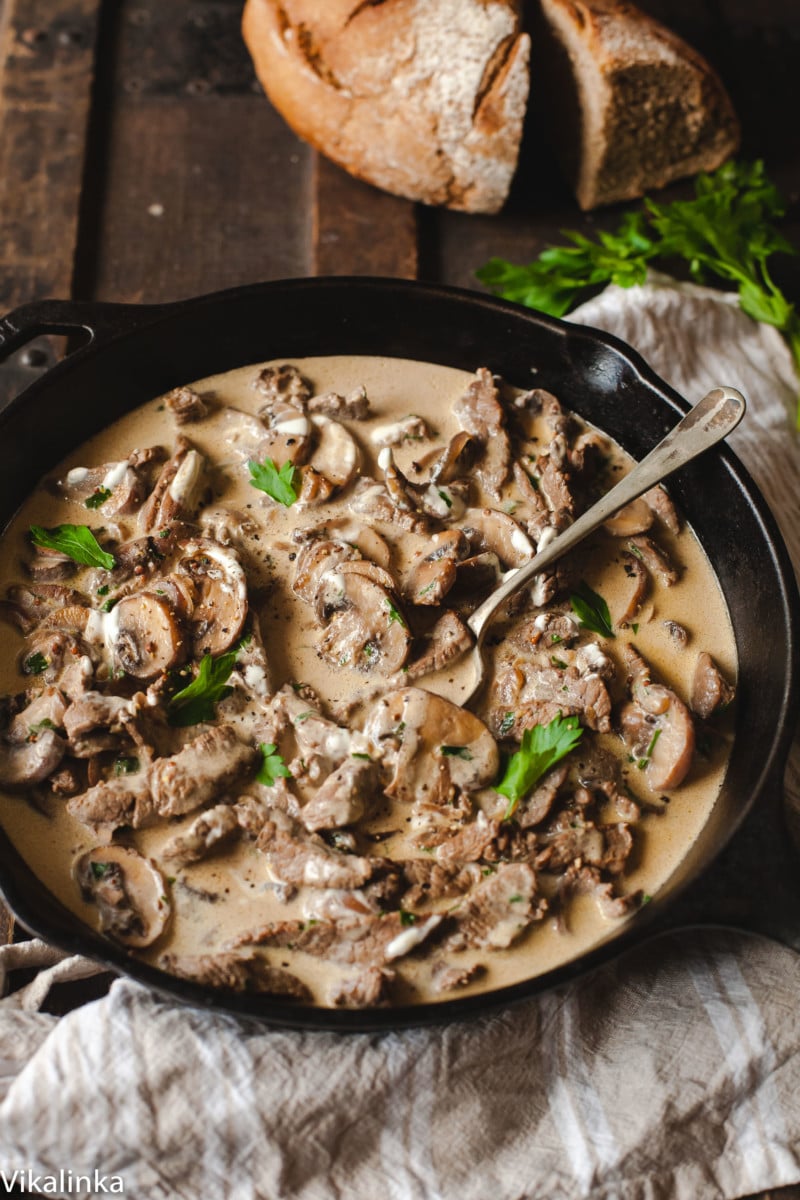 beef stroganoff in black pan on wooden table, loaf of bread in the background