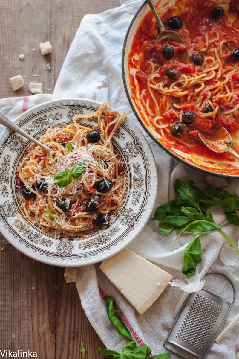 a bowl with spaghetti and black olives, a pan with spaghetti next to it, parmesan cheese and fresh basil. 