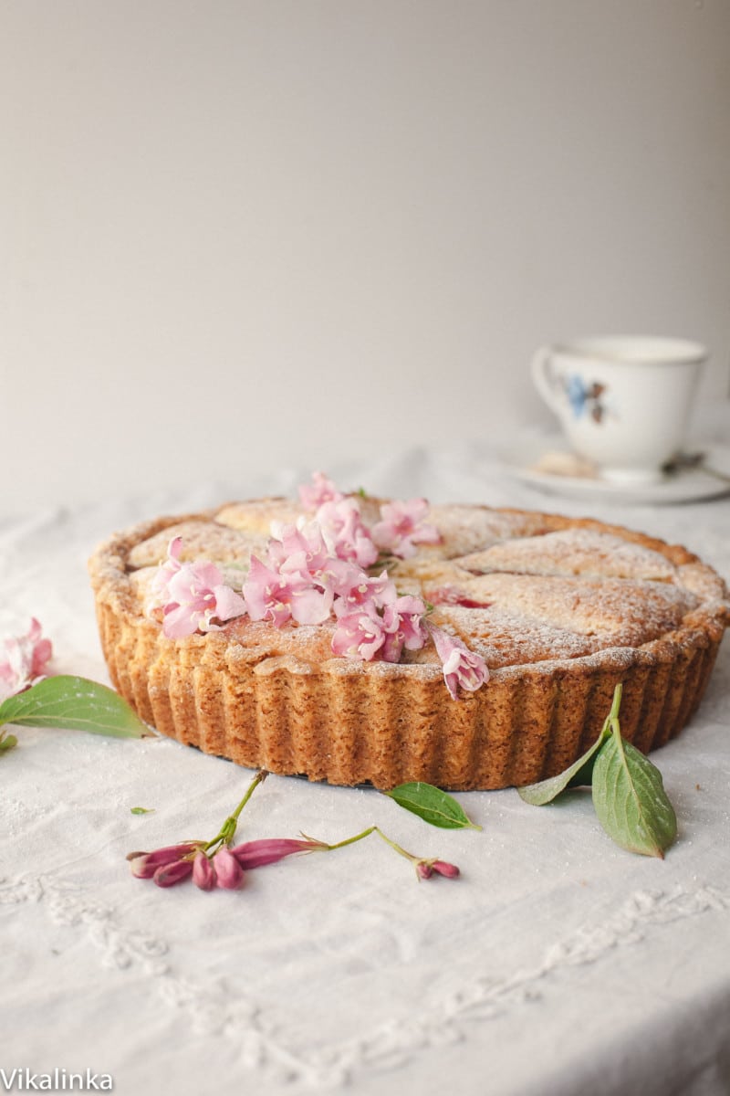 Bakewell tart decorated with pink flowers on a table covered with white tablecloth