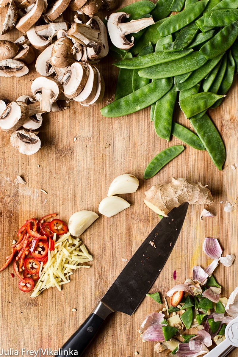 Sliced mushrooms, snow peas, sliced ginger and red chillies, garlic cloves and a knife on cutting board