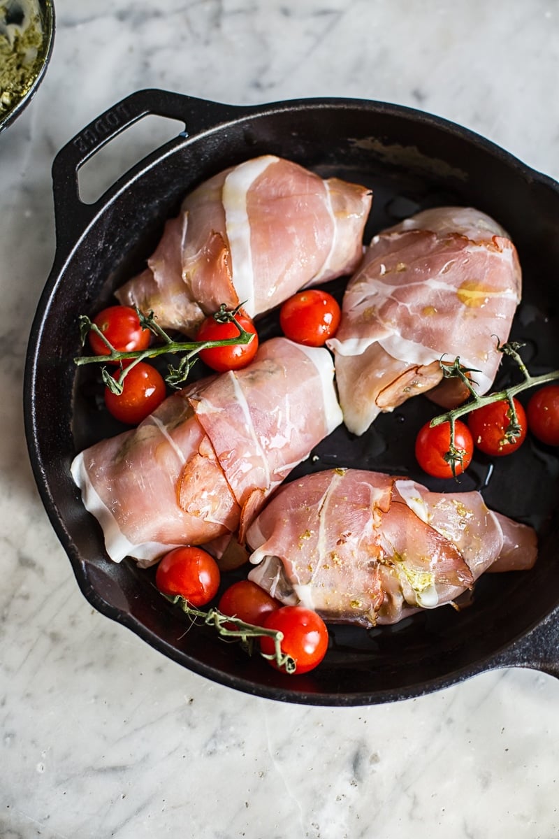Uncooked prosciutto wrapped pesto chicken parcels on a cast iron pan with cherry tomatoes