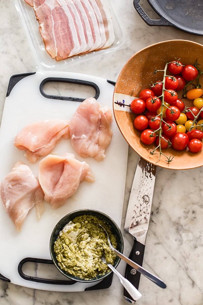 Ingredients for the recipe on a table, including pesto, tomatoes, chicken and proscuitto