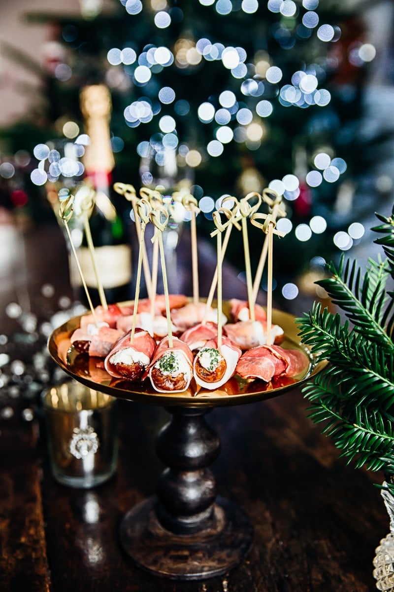 Herby Cheese Stuffed Dates Wrapped in Prosciutto on a platter with Christmas lights in the background