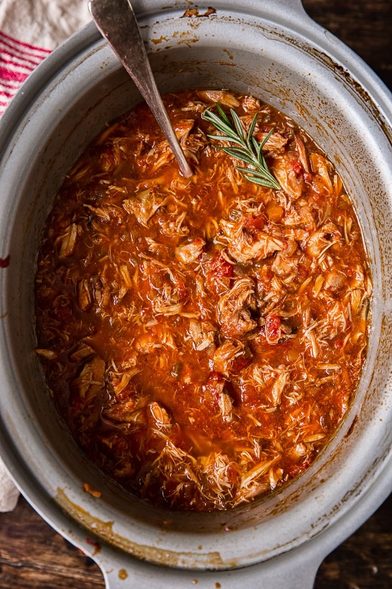 Top down of a slow cooker with the chicken ragu and a piece of rosemary