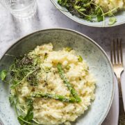top down view of two bowls of spring asparagus risotto with micro herbs visible, as well as a glass of water and a fork