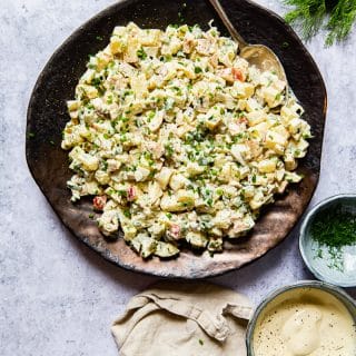 potato salad on a platter with a spoon, cream napkin, a bowl with salad dressing and a bowl with herbs, dill.