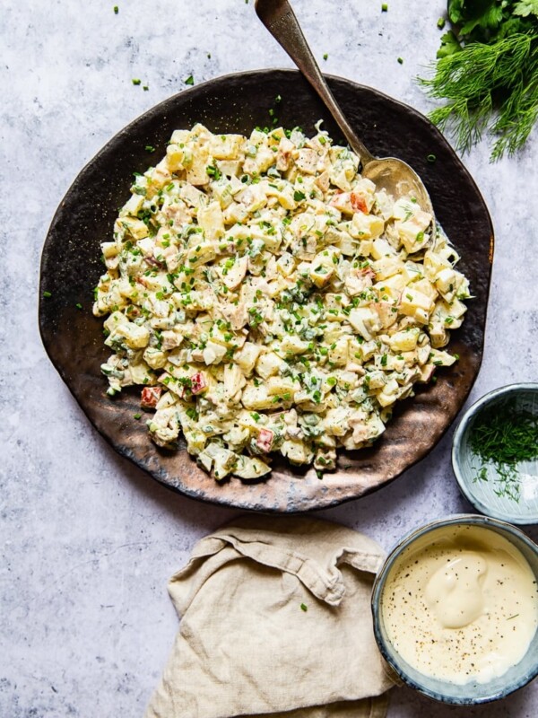 potato salad on a platter with a spoon, cream napkin, a bowl with salad dressing and a bowl with herbs, dill.