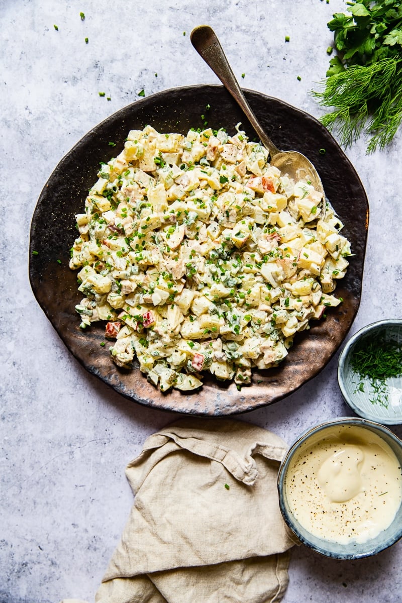 potato salad on a platter with a spoon, cream napkin, a bowl with salad dressing and a bowl with herbs, dill.