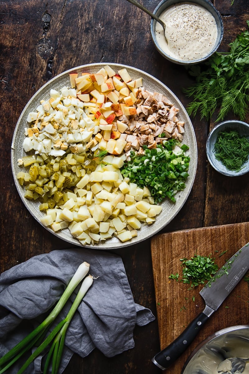 Russian salad ingredients on a platter, green onions, cutting board, herbs and salad dressing. 