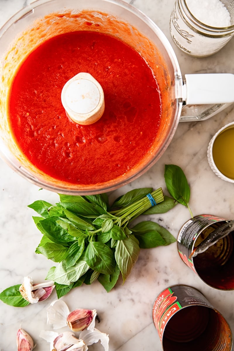 Food processor full of red sauce next to fresh basil and empty cans