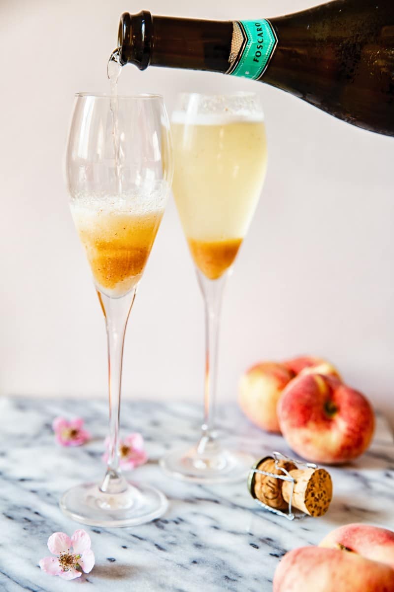 Two champagne glasses being filled with prosecco alongside peach