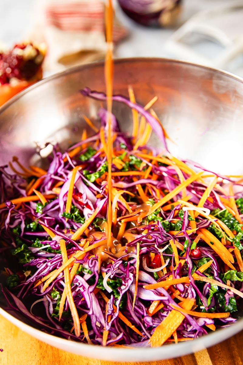 Sliced red cabbage, carrots, kale and pomegranate seeds in a bowl with dressing being poured over. 