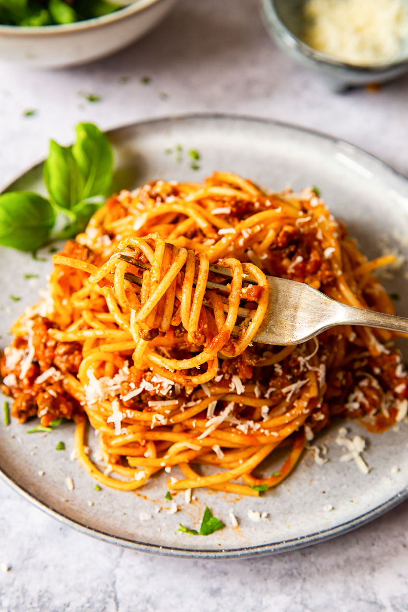 a fork with pasta coiled up with the pasta and meat sauce on a plate behind