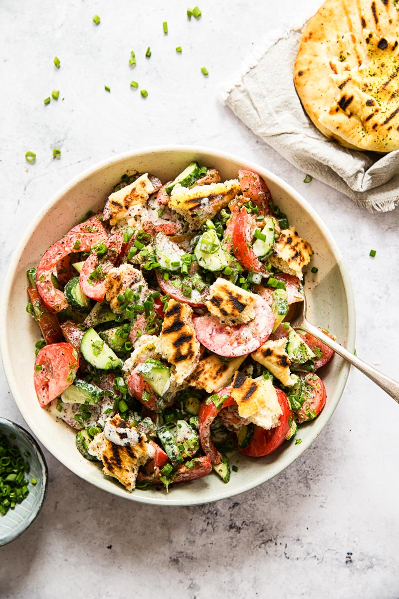 Overhead shot of fattoush salad in cream coloured bowl with a spoon, flatbread and a bowl with sliced green onions.