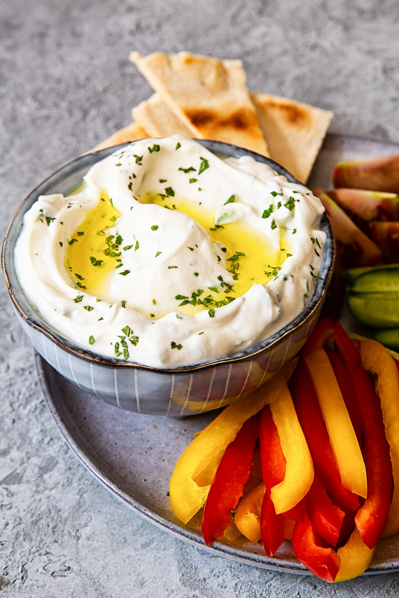 Greek tzatziki in blue bowl with cut up red and yellow peppers and pita bread