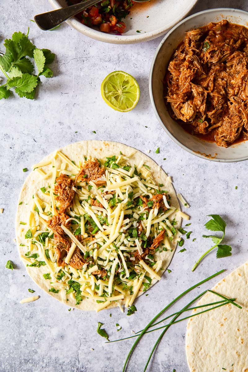 a tortilla topped with grated cheese and pulled pork and pulled pork in grey bowl