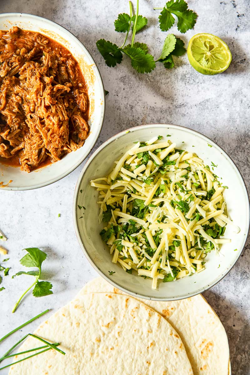 pulled pork and cheese and herb mixtures in two separate bowls on grey background. Flour tortillas next to them.