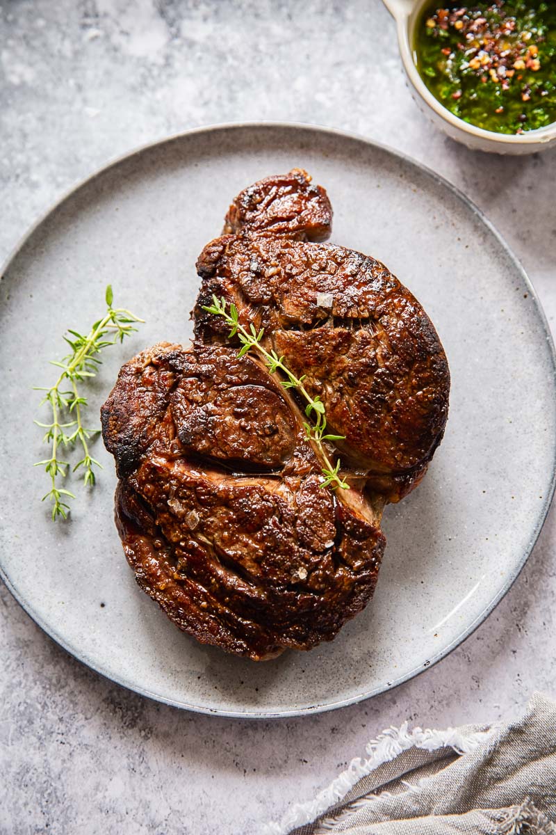 cooked ribeye steak on grey plate and a small dish of herb sauce near it