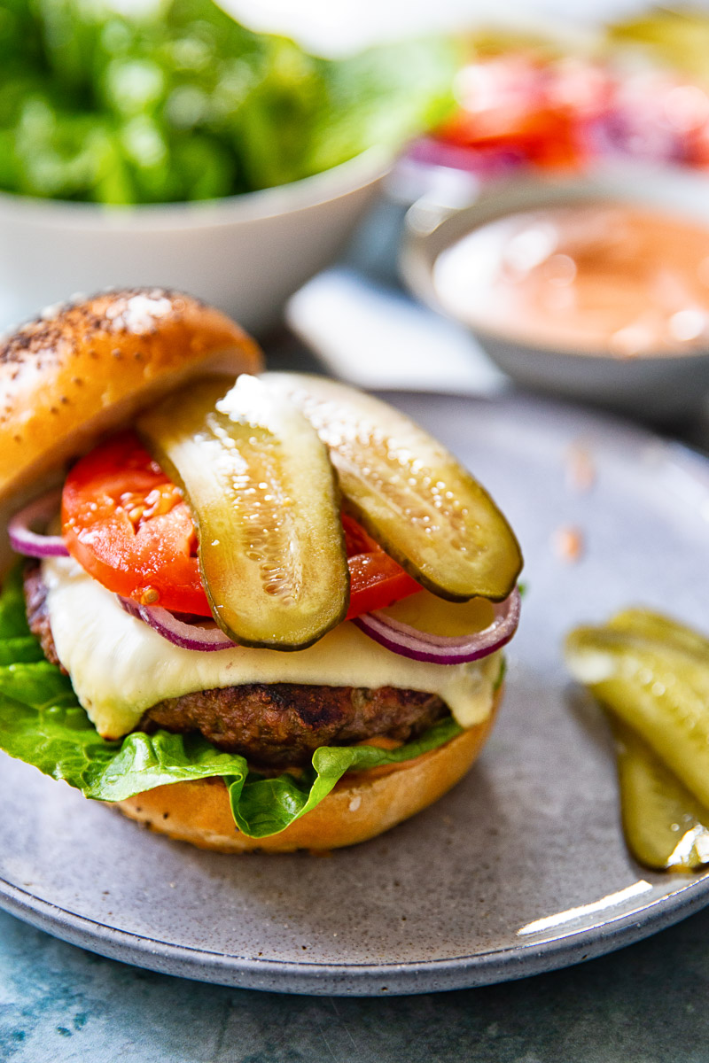 a burger topped with cheese, sliced onion, tomato and pickles resting on the bottom half of a bun