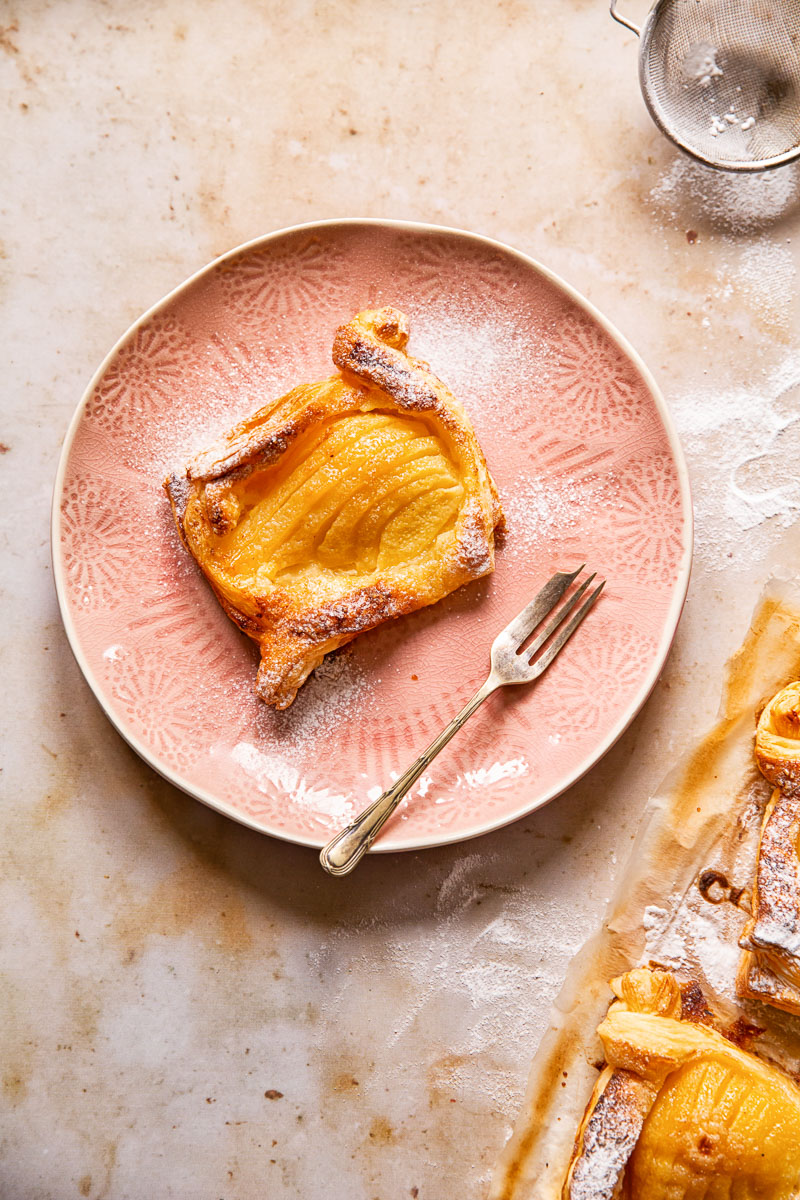 a pear tart on a pink plate, dessert fork, icing sugar in a sifter