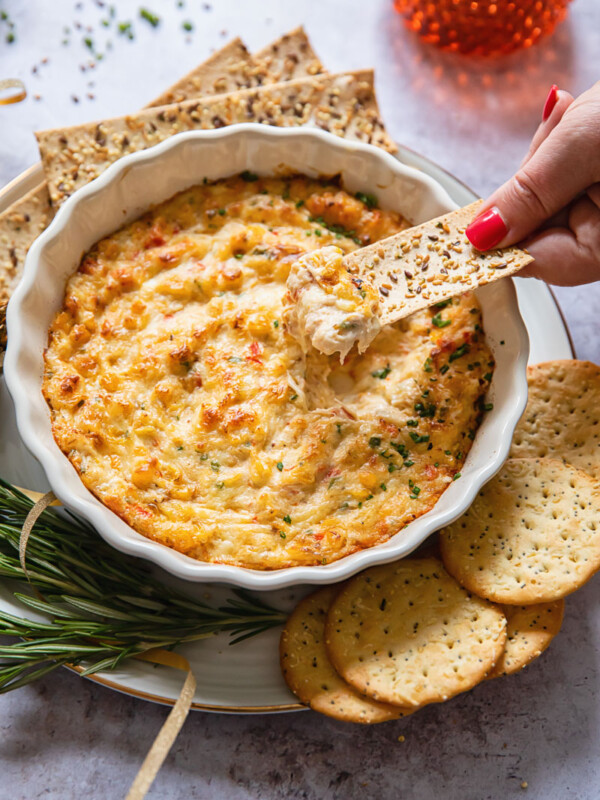 Holiday dip and a cracker being dipped into it, surrounded by crackers and fresh rosemary