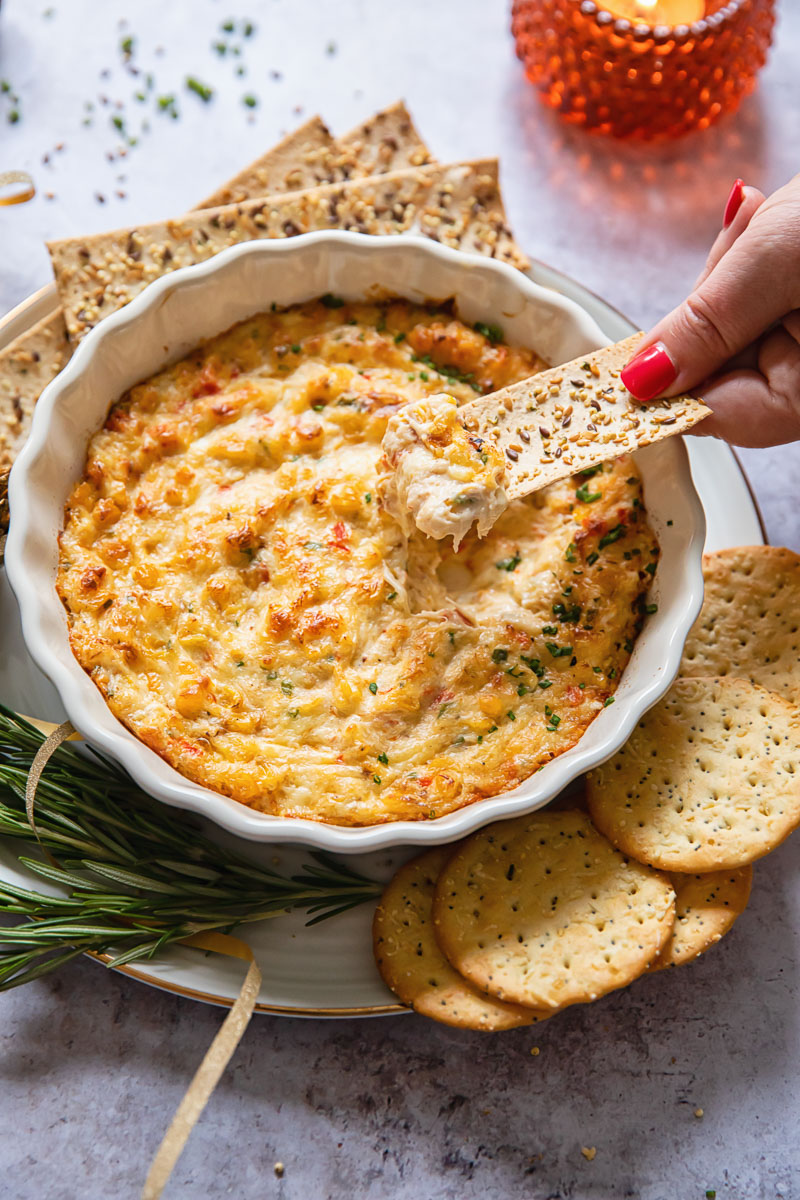 Holiday dip and a cracker being dipped into it, surrounded by crackers and fresh rosemary