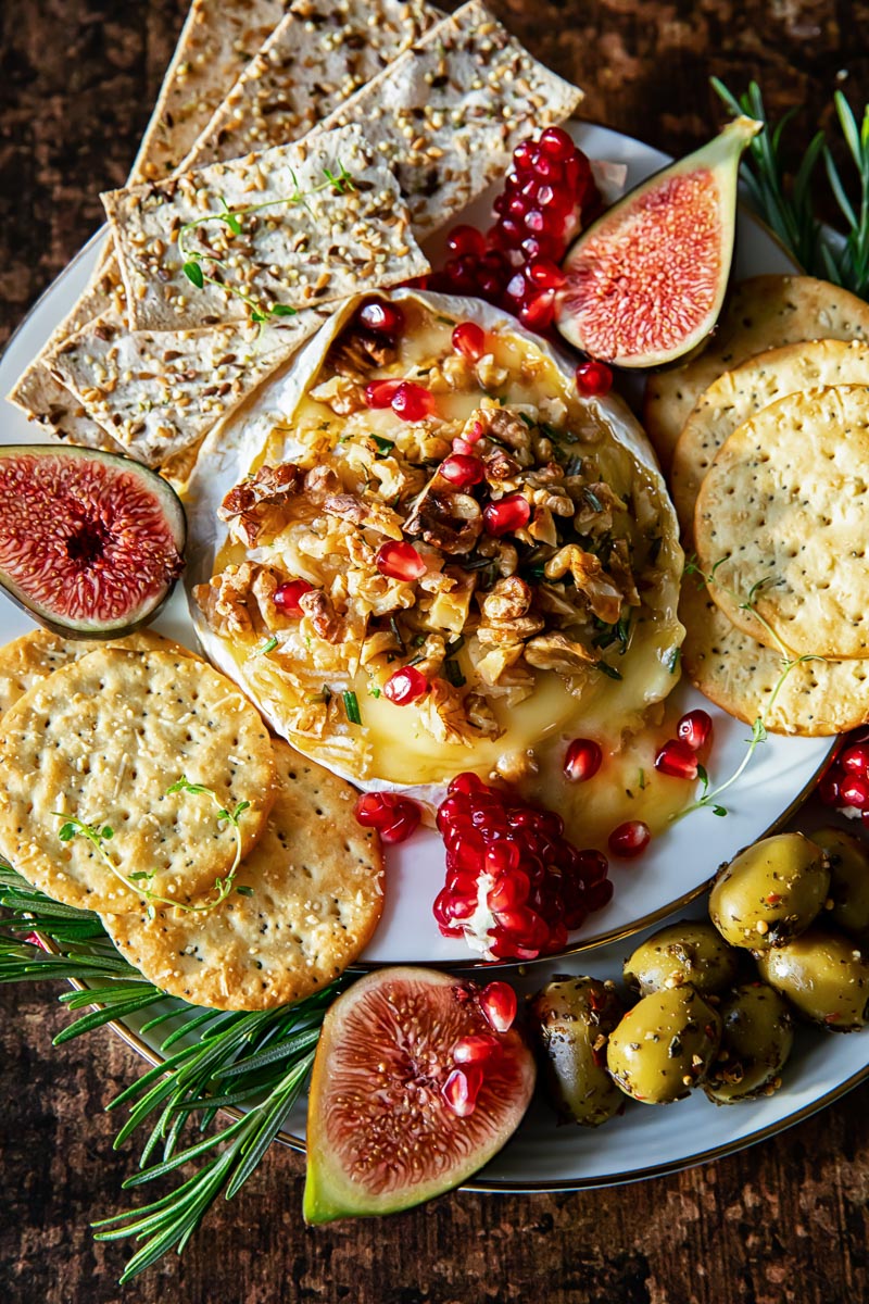 brie cheese topped with walnuts and pomegranate seed, surrounded by crackers, fresh figs and olives