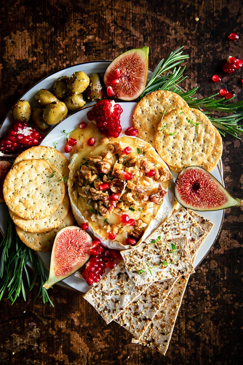 top down view of baked brie surrounded by crackers, olives, figs and pomegranate seeds