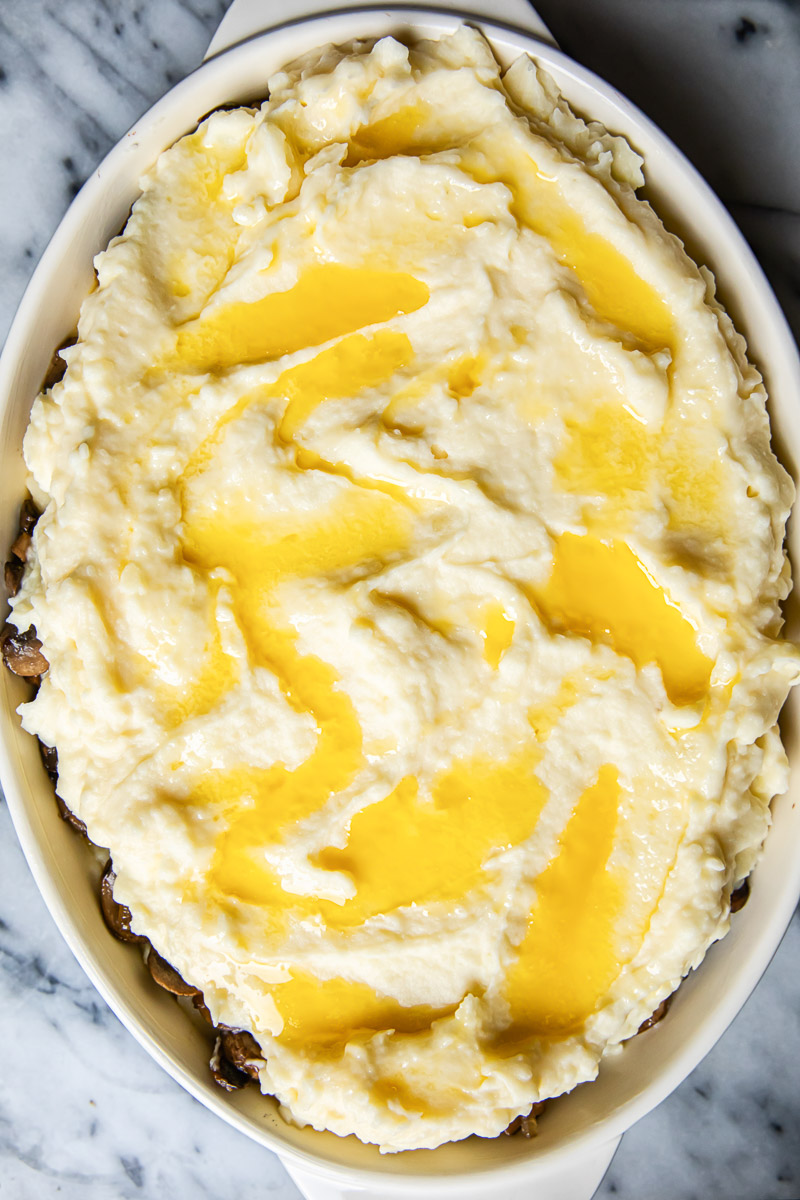 unbaked mashed potatoes in casserole dish with melted butter drizzled all over