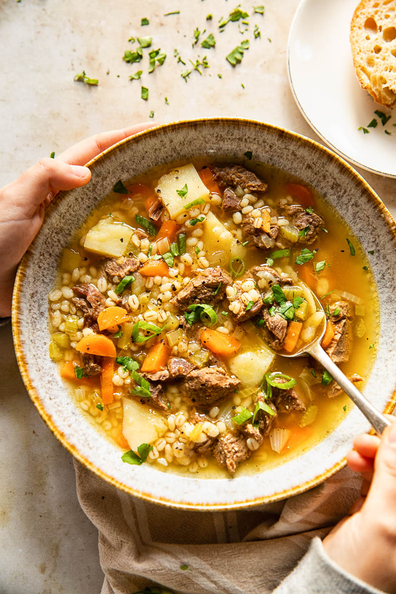 a bowl filled with beef, vegetable and barley soup, hands holding the bowl and a spoon.