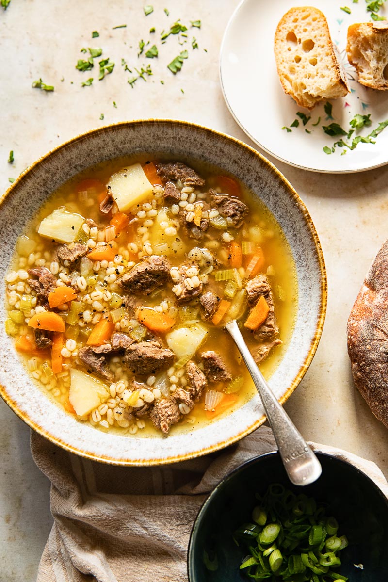 top down view of a bowl of soup with a spoon, a plate with slices of bread and a black bowl with chopped herbs