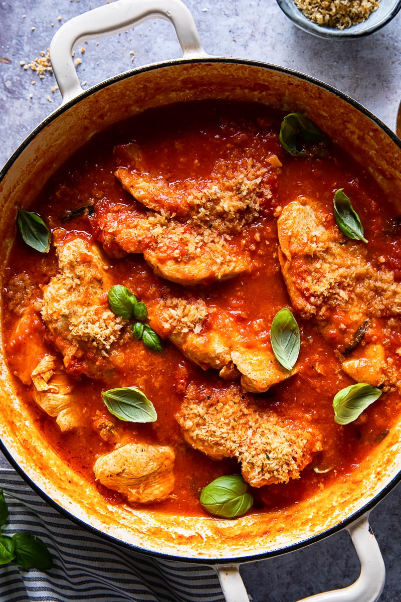 top down view of chicken in tomato sauce with fresh basil in a round cream coloured pan