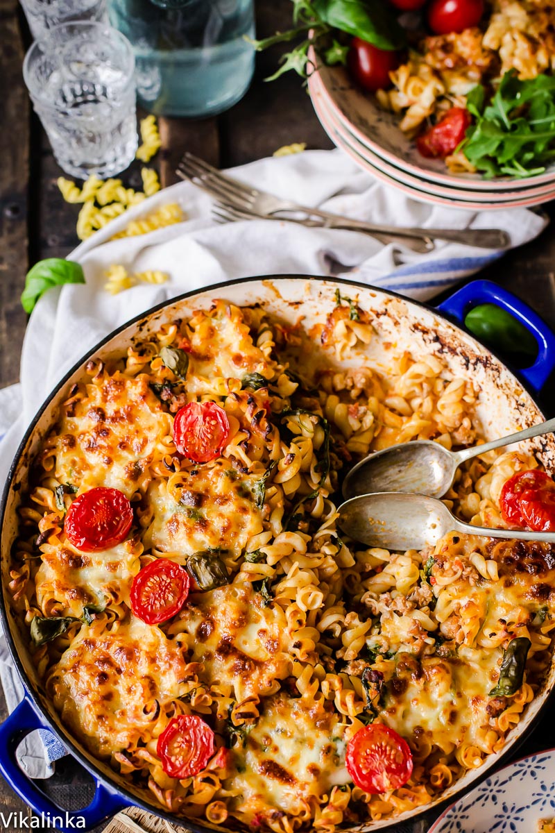 top down view of baked pasta topped with melted cheese and cherry tomatoes, bowls and glasses in the background. 