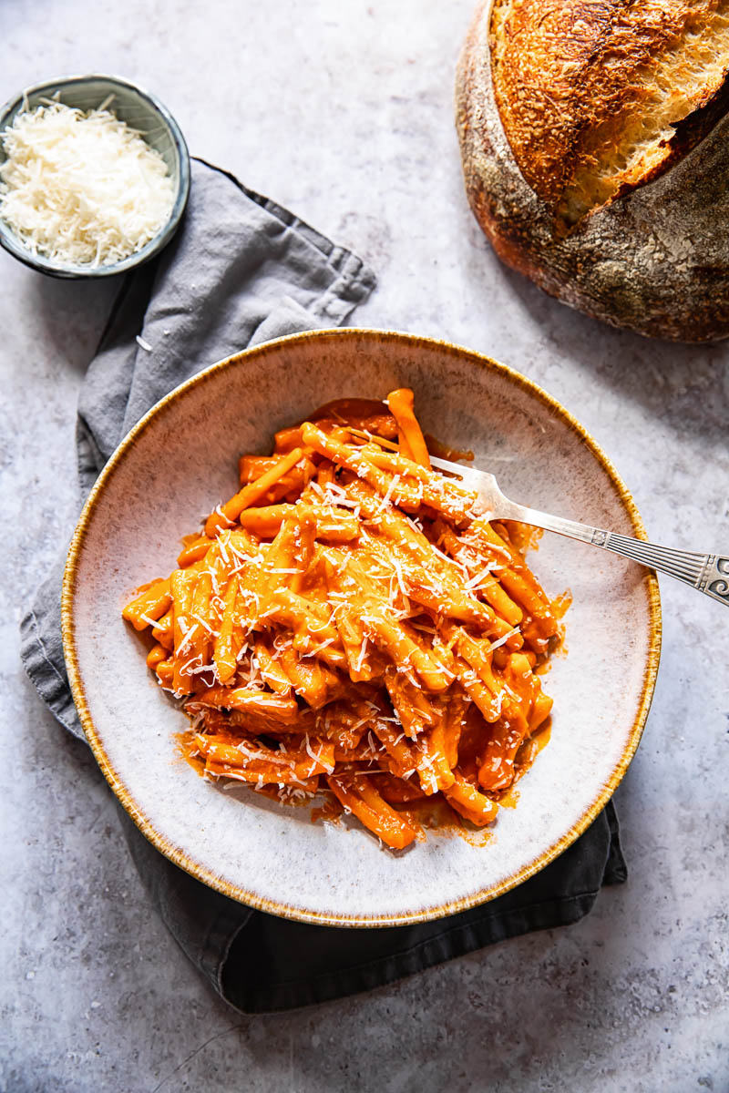 top down view of pasta alla vodka in a bowl with a fork, a load of bread and grated Parmesan in a small blue bowl next to it.