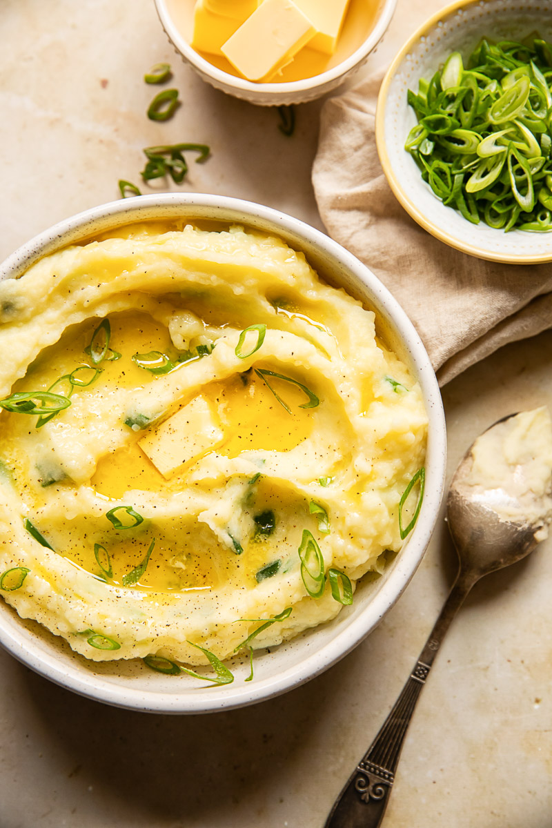 top down view of Irish Champ topped with melted butter, a spoon and a bowl with green onions and cubed butter next to it.