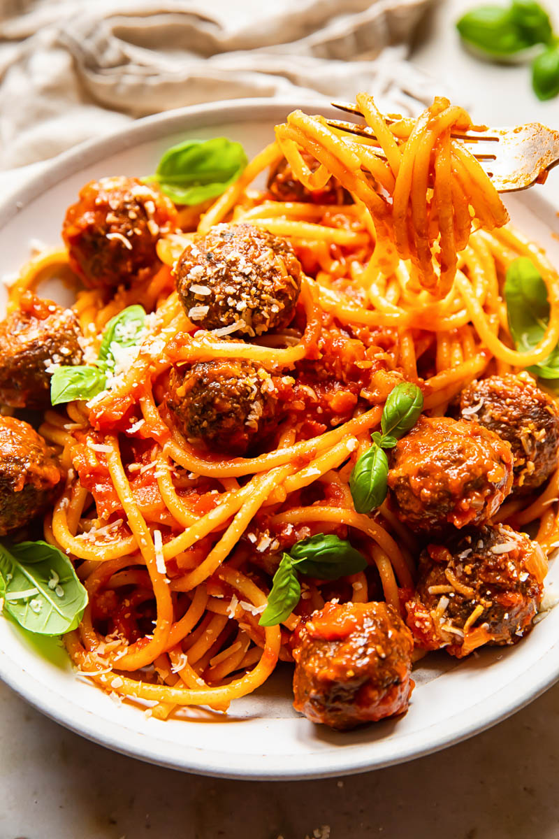 bucatini with meatballs in a cream coloured bowl, topped with fresh basil leaves