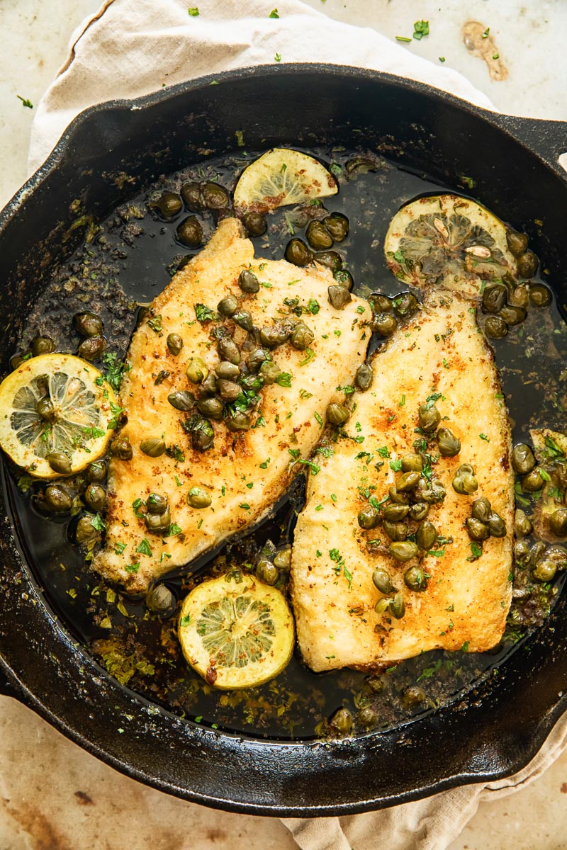 two sole fish topped with capers and lemon slices in cast iron pan