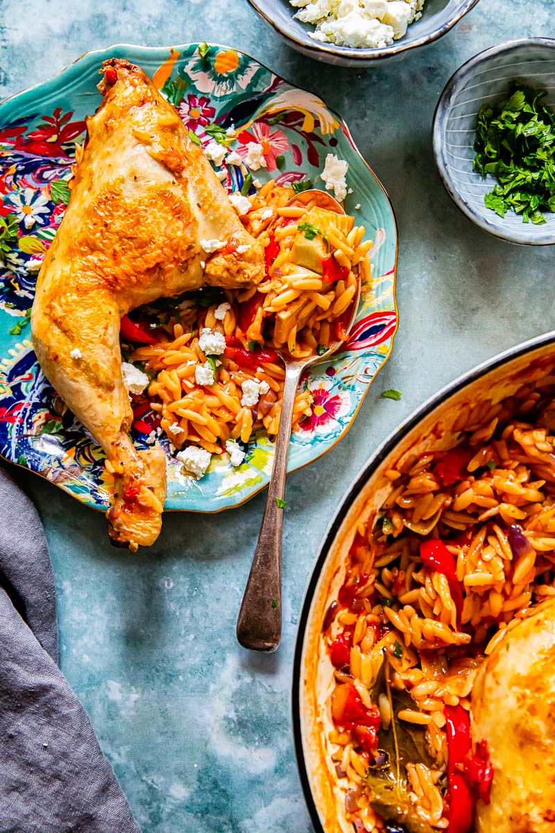 chicken leg and orzo on a blue plate, a pan with orzo, feta and parsley in small bowls. 