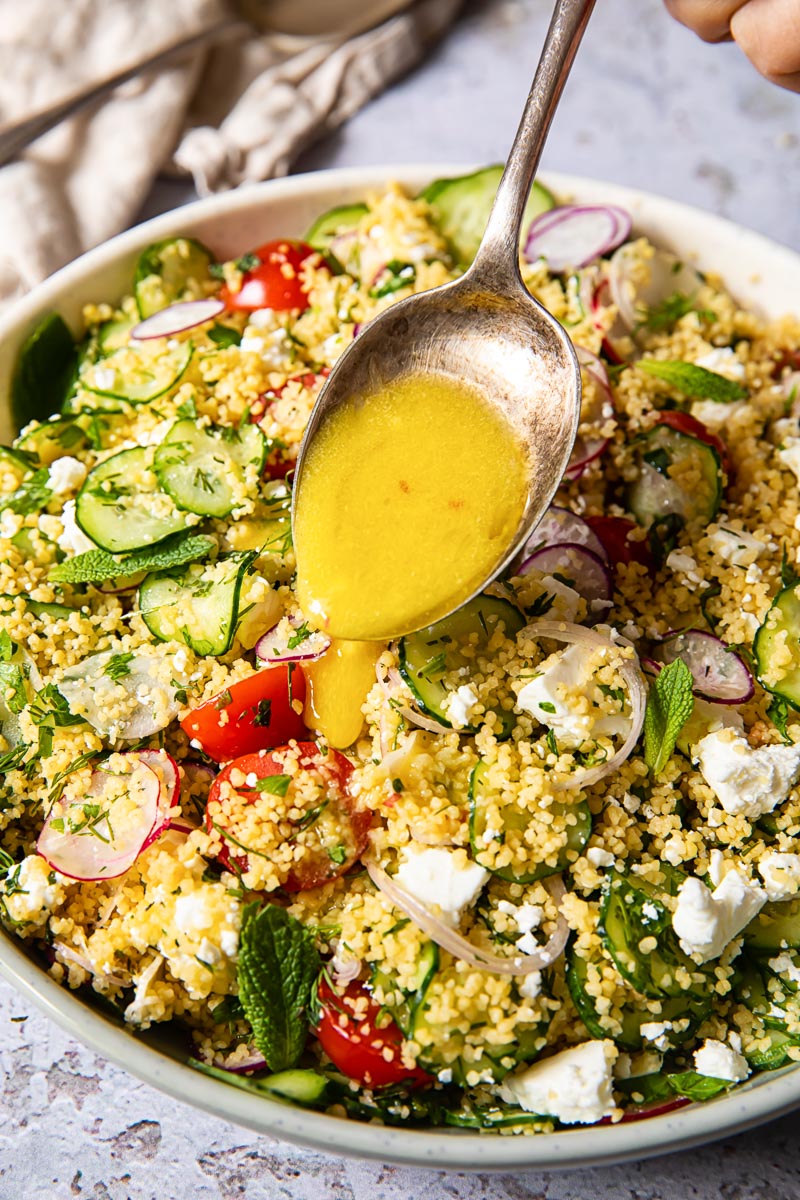 a spoon with a lemon salad dressing being drizzled over couscous salad