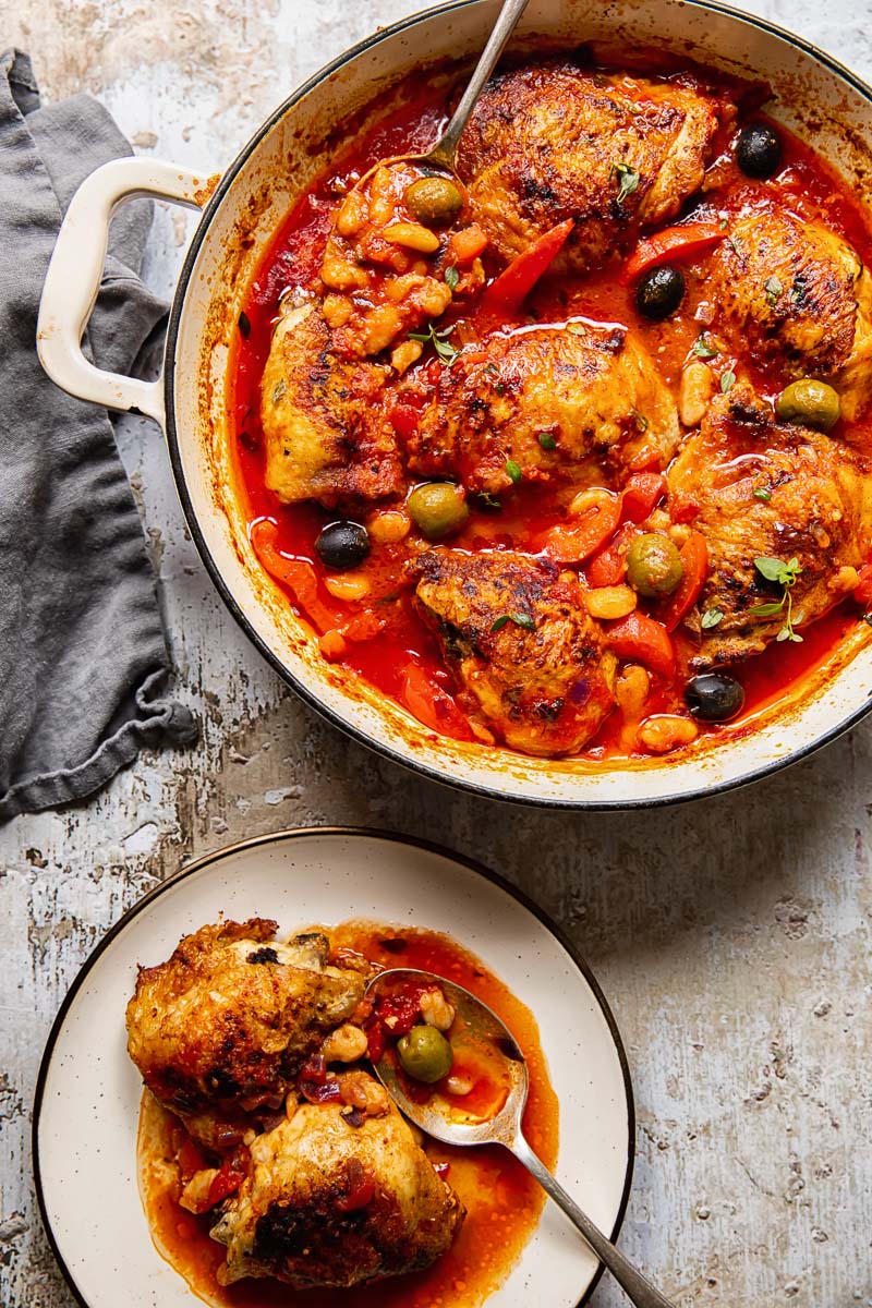 Chicken thighs with red peppers and olives in a tomato sauce in a pan, a plate with chicken and beans with a spoon.