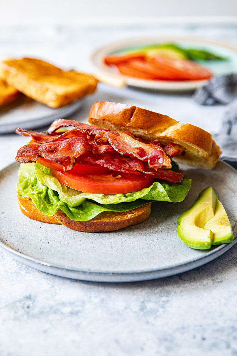 side view of bacon, lettuce, tomato sandwich on grey plate, avocado next to it
