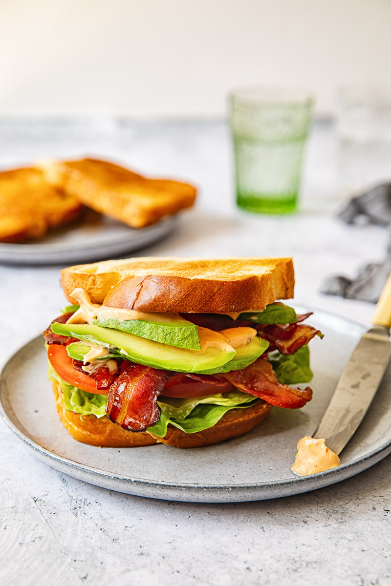 sandwich on grey plate, butter knife on the plate, bread sliced and green water glass in the background.