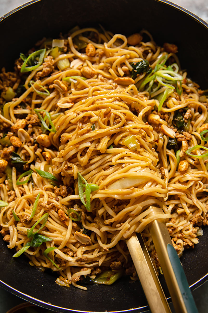 noodles in a black wok with tongs holding the noodles
