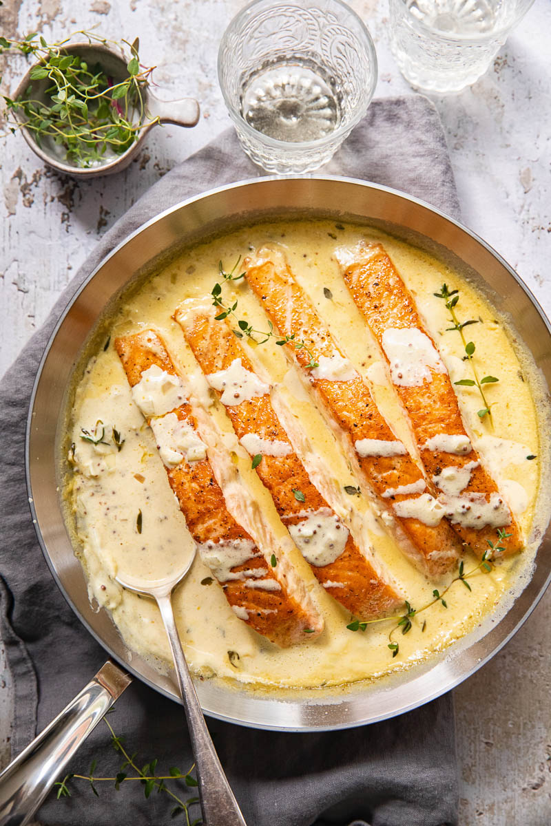 top down view of salmon pieces in cream sauce in a pan, water glasses, herbs on the table.