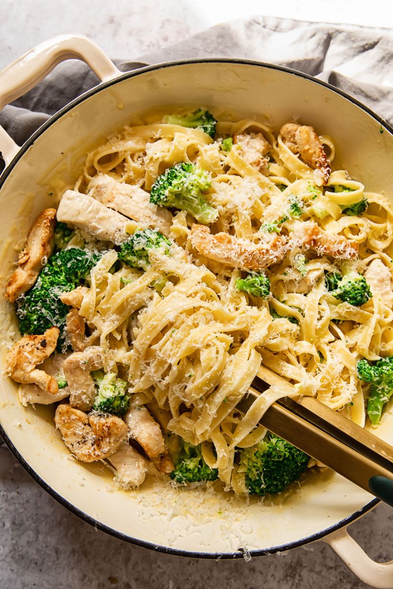 top down view of chicken broccoli alfredo fettuccine in a pan with tongs in it