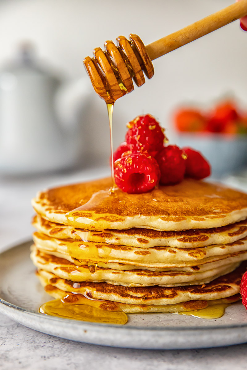 side view of a stack of pancakes and raspberries being drizzled with maple syrup