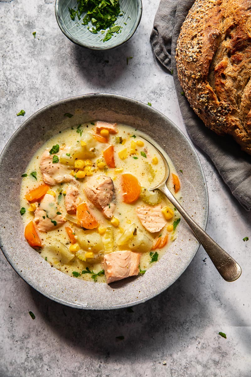 salmon chowder in a grey bowl with a spoon in it, a loaf of bread in the corner