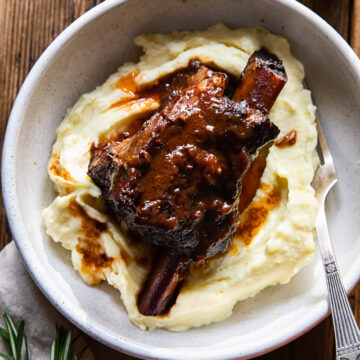 top down view of a braised short rib on top of mashed potatoes in a bowl, a glass of wine next to it