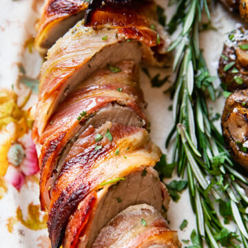 sliced pork tenderloin wrapped in bacon on a platter with rosemary sprig next to it